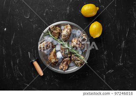 Fresh closed oysters, ice and lemon on a round metal plate, black stone textured background. Top view with copy space. Close-up shot. Fresh closed oysters, ice and lemon on a round metal plate, black stone textured background. Top view with copy space. Close-up shot. 97246018