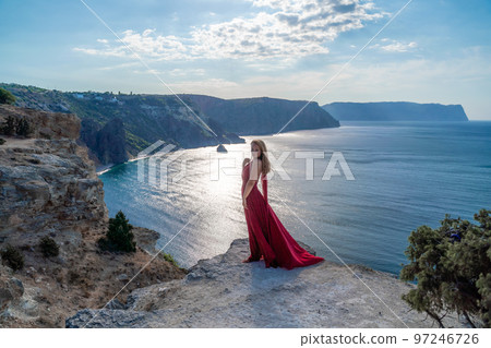 A girl with flowing hair in a long red dress stands on a rock above the sea. The stone can be seen in the sea. Sunny path to the sea from the sun. 97246726