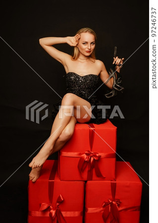 Woman gifts christmas. Beautiful sexy young blonde in a black dress sits on three big red gifts on a black background. Christmas photo, gifts Woman gifts christmas. Beautiful sexy young blonde in a black dress sits on three big red gifts on a black background. Christmas photo, gifts 97246737