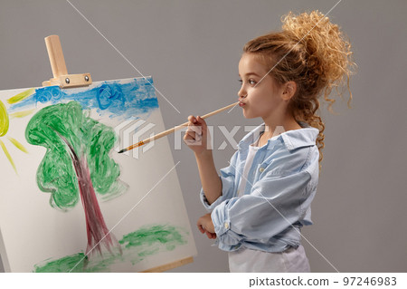 Charming school girl is painting with a watercolor brush on an easel, standing on a gray background. 97246983