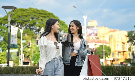 Two stylish young women with shopping bag standing in city avenue with blurred cityscape and beautiful blue sky in background Two stylish young women with shopping bag standing in city avenue with blurred cityscape and beautiful blue sky in background 97247113