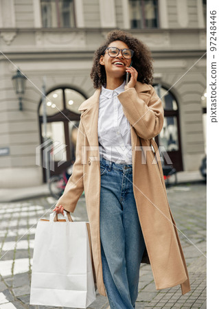 Smiling afro american woman after shopping with paper bags and phone while walking in city Smiling afro american woman after shopping with paper bags and phone while walking in city 97248446