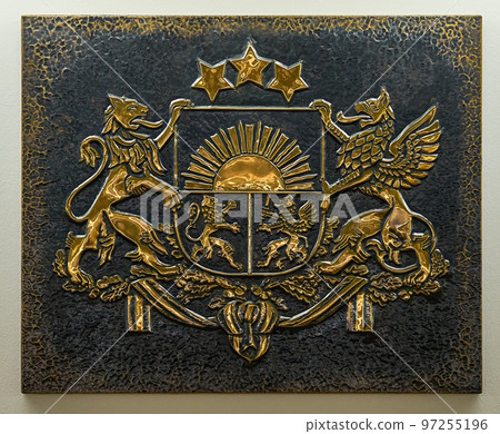 Closeup of gold color coat of arms of Latvia embossed on a relief dark metal plate. 97255196