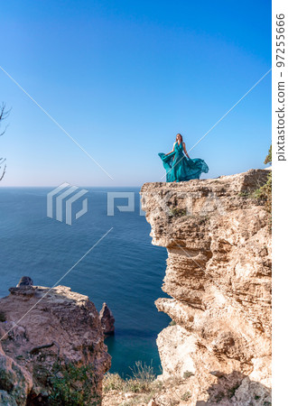 A girl with loose hair in a long mint dress descends the stairs between the yellow rocks overlooking the sea. A rock can be seen in the sea. Sunny path on the sea from the rising sun A girl with loose hair in a long mint dress descends the stairs between the yellow rocks overlooking the sea. A rock can be seen in the sea. Sunny path on the sea from the rising sun 97255666