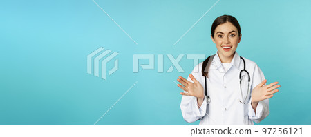 Happy and excited smiling young woman looking with excitement and surprised, standing in white lab coat and stethoscope, blue background Happy and excited smiling young woman looking with excitement and surprised, standing in white lab coat and stethoscope, blue background 97256121