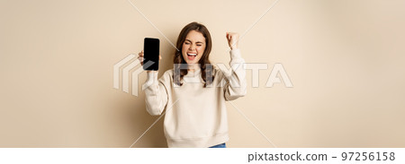 Happy woman winning, showing smartphone screen and shouting of joy, fist pump, standing over beige background Happy woman winning, showing smartphone screen and shouting of joy, fist pump, standing over beige background 97256158