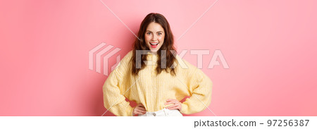 Portrait of sassy and confident female model, holding hands on waist and smiling, saying yes with self-assured face, standing against pink background 97256387