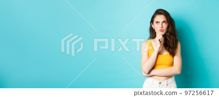 Portrait of attractive girl in summer outfit thinking, making decision, frowning and looking at upper left corner with pensive face, standing over blue background 97256617