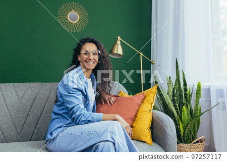 Portrait of cheerful hispanic woman at home sitting on couch wearing glasses smiling and looking at camera. 97257117