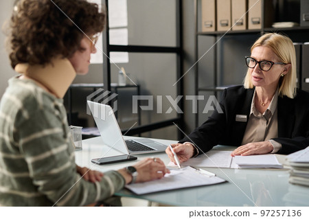 Portrait of female insurance broker consulting injured young woman in office after accident and pointing at legal forms 97257136