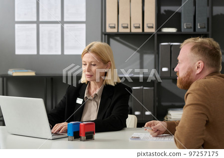 Portrait of mature woman working in services office and using laptop while consulting male client 97257175