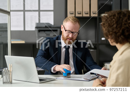 Portrait of bearded man stamping application form while working with female visitor in agency office 97257211