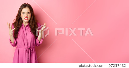 Angry and furious young woman shaking hands, looks frustrated and outraged, arguing, complaining on something, standing against pink background 97257482
