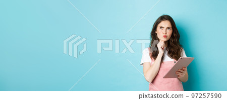 Image of thoughtful female student looking at upper left corner and thinking, holding digital tablet, making hard choice, standing over blue background 97257590