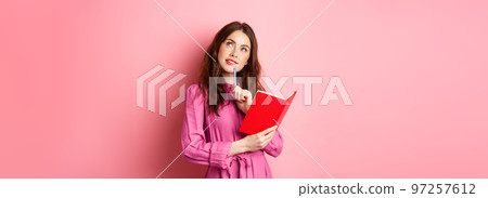 Beautiful young woman look thoughtful, writing in notebook, holding planner or diary, plan her schedule, standing against pink background 97257612