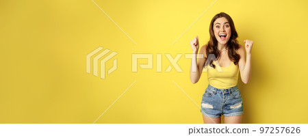 Enthusiastic young woman shouting and cheering, rejoicing, raising hands up and screaming in joy, chanting, standing over yellow background Enthusiastic young woman shouting and cheering, rejoicing, raising hands up and screaming in joy, chanting, standing over yellow background 97257626