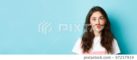 Close up portrait of funny silly girl playing, making squinting face and moustache with hair strand above lips, standing over blue background 97257919