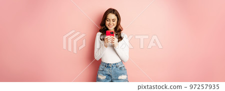 Smiling brunette girl using smartphone, texting message, order online on mobile phone app, standing over pink background Smiling brunette girl using smartphone, texting message, order online on mobile phone app, standing over pink background 97257935