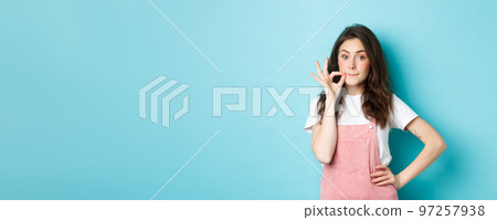 Cute glamour girl making lips zipper gesture, lock her mouth, promise keep secret, standing against blue background Cute glamour girl making lips zipper gesture, lock her mouth, promise keep secret, standing against blue background 97257938