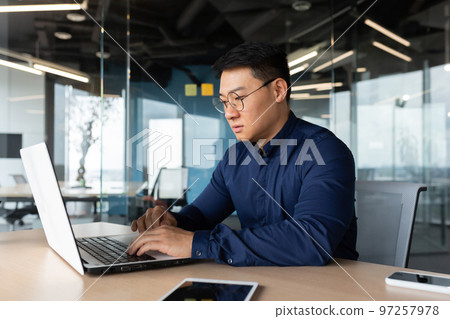 Serious and thoughtful businessman working inside office sitting at table using laptop at work, mature asian boss in shirt thinking and typing on keyboard. Serious and thoughtful businessman working inside office sitting at table using laptop at work, mature asian boss in shirt thinking and typing on keyboard. 97257978