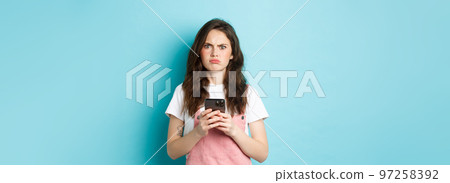 Disappointed girl frowning and grimacing displeased after using smartphone app, holding phone and express dislike, standing over blue background 97258392