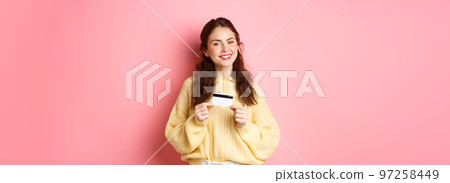 Beautiful female model thinking of shopping, holding plastic credit card and smiling, standing against pink spring background Beautiful female model thinking of shopping, holding plastic credit card and smiling, standing against pink spring background 97258449