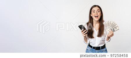 Online microcredit loans and banking concept. Happy woman holding mobile phone and money, smiling and laughing, standing over white background Online microcredit loans and banking concept. Happy woman holding mobile phone and money, smiling and laughing, standing over white background 97258458