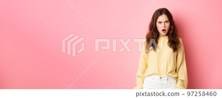 Shocked and confused brunette woman gasping, frowning and looking offended at camera, feeling insulted or displeased, standing over pink background 97258460