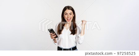 Enthusiastic woman winning on mobile phone, rejoicing and screaming of joy while using smartphone app, standing over white background 97258525