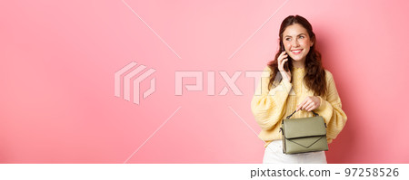 Beautiful and stylish lady having phone call, smiling while talking with someone on cellphone, holding purse, standing against pink background Beautiful and stylish lady having phone call, smiling while talking with someone on cellphone, holding purse, standing against pink background 97258526