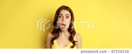 Close up portrait of stylish brunette girl looking amazed, wow face, showing excitement and surprise, impressed by something, standing over yellow background Close up portrait of stylish brunette girl looking amazed, wow face, showing excitement and surprise, impressed by something, standing over yellow background 97258538