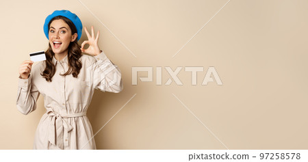 Concept of shopping and sale. Happy young woman in stylish clothes, showing credit card store discount and okay, satisfied, like and approve smth, standing over beige background Concept of shopping and sale. Happy young woman in stylish clothes, showing credit card store discount and okay, satisfied, like and approve smth, standing over beige background 97258578