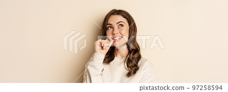 Cute modern woman thinking, daydreaming and smiling, looking up thoughtful, standing over beige background Cute modern woman thinking, daydreaming and smiling, looking up thoughtful, standing over beige background 97258594