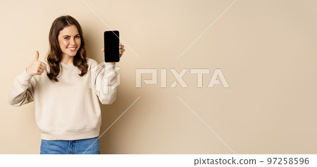 Attractive smiling woman showing thumbs up and smartphone screen, standing over beige background Attractive smiling woman showing thumbs up and smartphone screen, standing over beige background 97258596