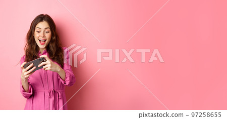 Technology concept. Beautiful modern girl playing racing video game on smartphone, tap mobile screen and smiling happy, pink background 97258655