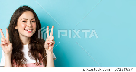 Portrait of cute smiling girl showing peace signs and looking happy at camera, standing in spring outfit against blue background Portrait of cute smiling girl showing peace signs and looking happy at camera, standing in spring outfit against blue background 97258657