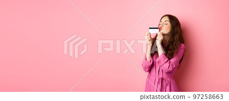 Image of young beautiful woman kissing her credit card, going on shopping, wasting money in stores, standing against pink background Image of young beautiful woman kissing her credit card, going on shopping, wasting money in stores, standing against pink background 97258663