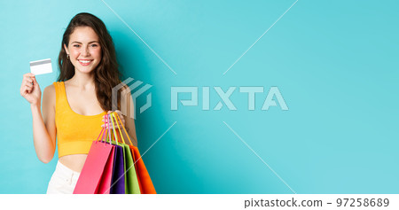 Stylish attractive woman showing plastic credit card she used to pay for new clothes, holding shopping bags with goods, standing over blue background 97258689