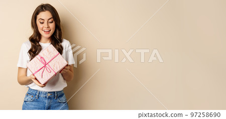 Celebration and holidays concept. Happy young woman holding gift wrapped in pink box, receive present, looking amazed and surprised, standing over beige background 97258690