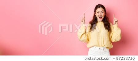 Cheerful young woman showing advertisement, pointing fingers up at promotional text and smiling excited, standing against pink background Cheerful young woman showing advertisement, pointing fingers up at promotional text and smiling excited, standing against pink background 97258730