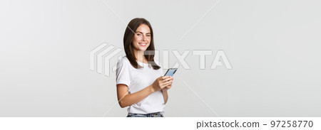Attractive young brunette woman using mobile phone and smiling at camera Attractive young brunette woman using mobile phone and smiling at camera 97258770