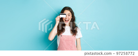 Excited girl holding plastic credit card near eye and look amazed at camera, buying things in store, going shopping, wasting money on bank account, standing over blue background Excited girl holding plastic credit card near eye and look amazed at camera, buying things in store, going shopping, wasting money on bank account, standing over blue background 97258772