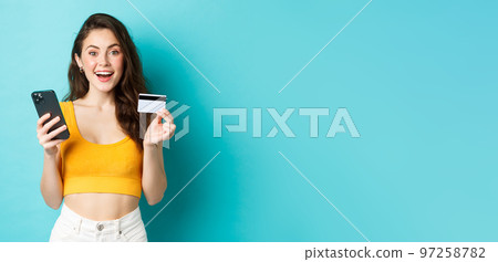 Young happy woman making online order, shopping with credit card and smartphone, look amazed at camera, standing over blue background Young happy woman making online order, shopping with credit card and smartphone, look amazed at camera, standing over blue background 97258782