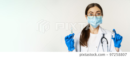 Covid-19 vaccination and healthcare concept. Young doctor in medical mask showing syringe and vaccine from coronavirus omicron variant, standing over white background Covid-19 vaccination and healthcare concept. Young doctor in medical mask showing syringe and vaccine from coronavirus omicron variant, standing over white background 97258807