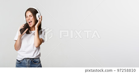 Attractive happy woman playing karaoke app, singing into smartphone, wearing wireless headphones 97258810