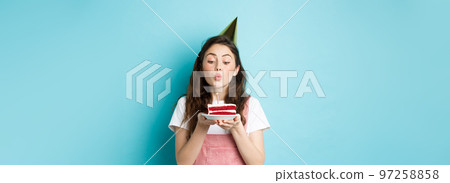 Holidays and celebration. Excited woman celebrating birthday, blowing candle on cake, wearing party cake and having fun, standing over blue background Holidays and celebration. Excited woman celebrating birthday, blowing candle on cake, wearing party cake and having fun, standing over blue background 97258858