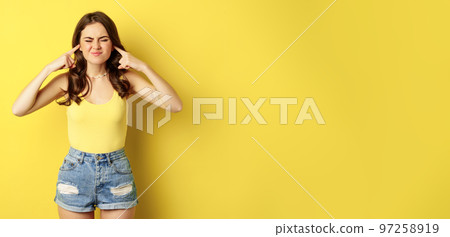 Young stylish woman feeling discomfort from loud noise sound, shut ears with fingers and complaining, standing over yellow background Young stylish woman feeling discomfort from loud noise sound, shut ears with fingers and complaining, standing over yellow background 97258919