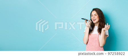 Portrait of smiling woman holding phone near lips and talking, using app translator on smartphone or recording voice message, speaking with speakerphone, standing over blue background Portrait of smiling woman holding phone near lips and talking, using app translator on smartphone or recording voice message, speaking with speakerphone, standing over blue background 97258922