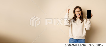 Happy woman winning, showing smartphone screen and shouting of joy, fist pump, standing over beige background Happy woman winning, showing smartphone screen and shouting of joy, fist pump, standing over beige background 97259057