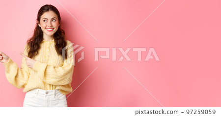 Young happy woman smiling, pointing and looking aside at left copyspace, showing advertisement, standing against pink spring background Young happy woman smiling, pointing and looking aside at left copyspace, showing advertisement, standing against pink spring background 97259059